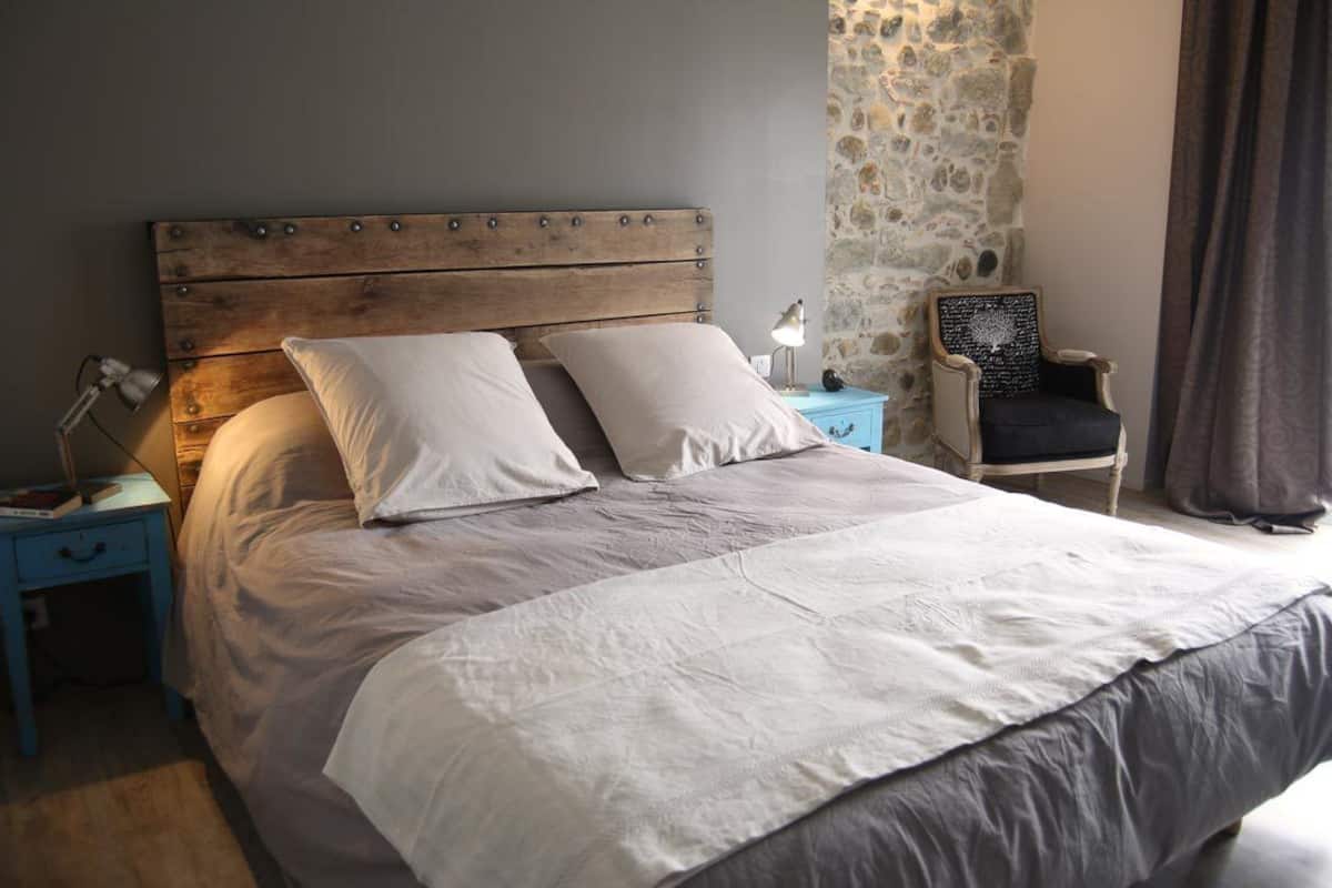 Un dormitorio con cabecera de madera, dos mesitas de noche, una silla y pared de piedra.