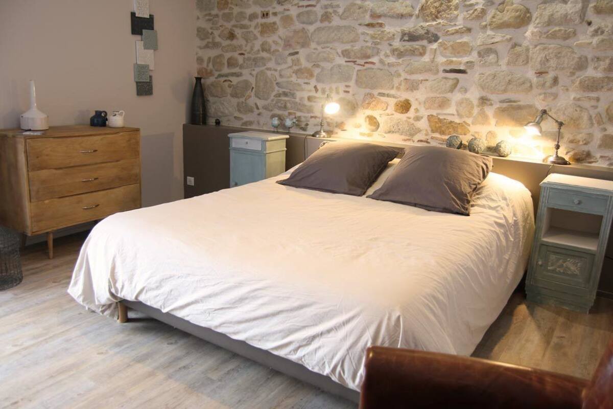 Un dormitorio con pared de piedra, una cama con dos almohadas, un aparador de madera y una mesita de noche con una lámpara.