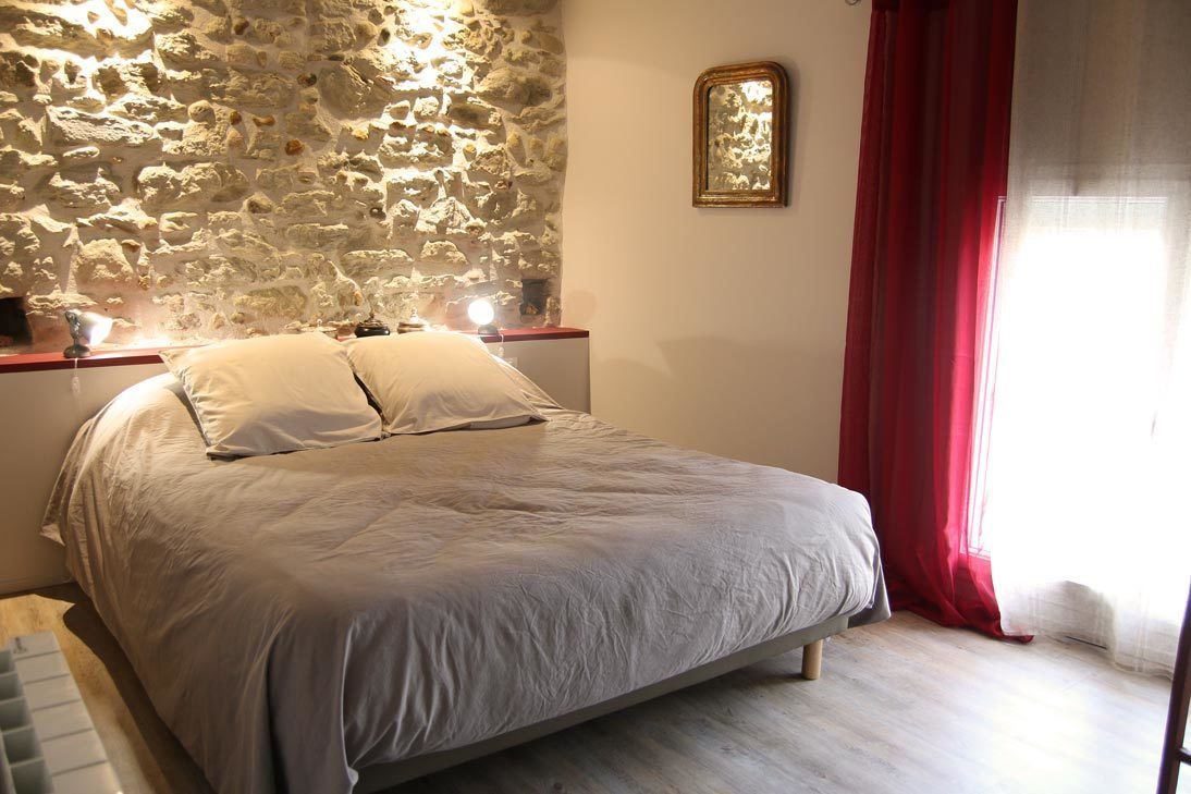 Une chambre avec un mur en pierre, un lit recouvert de linge de lit blanc, une table de chevet avec une lampe et une fenêtre ornée de rideaux rouges.