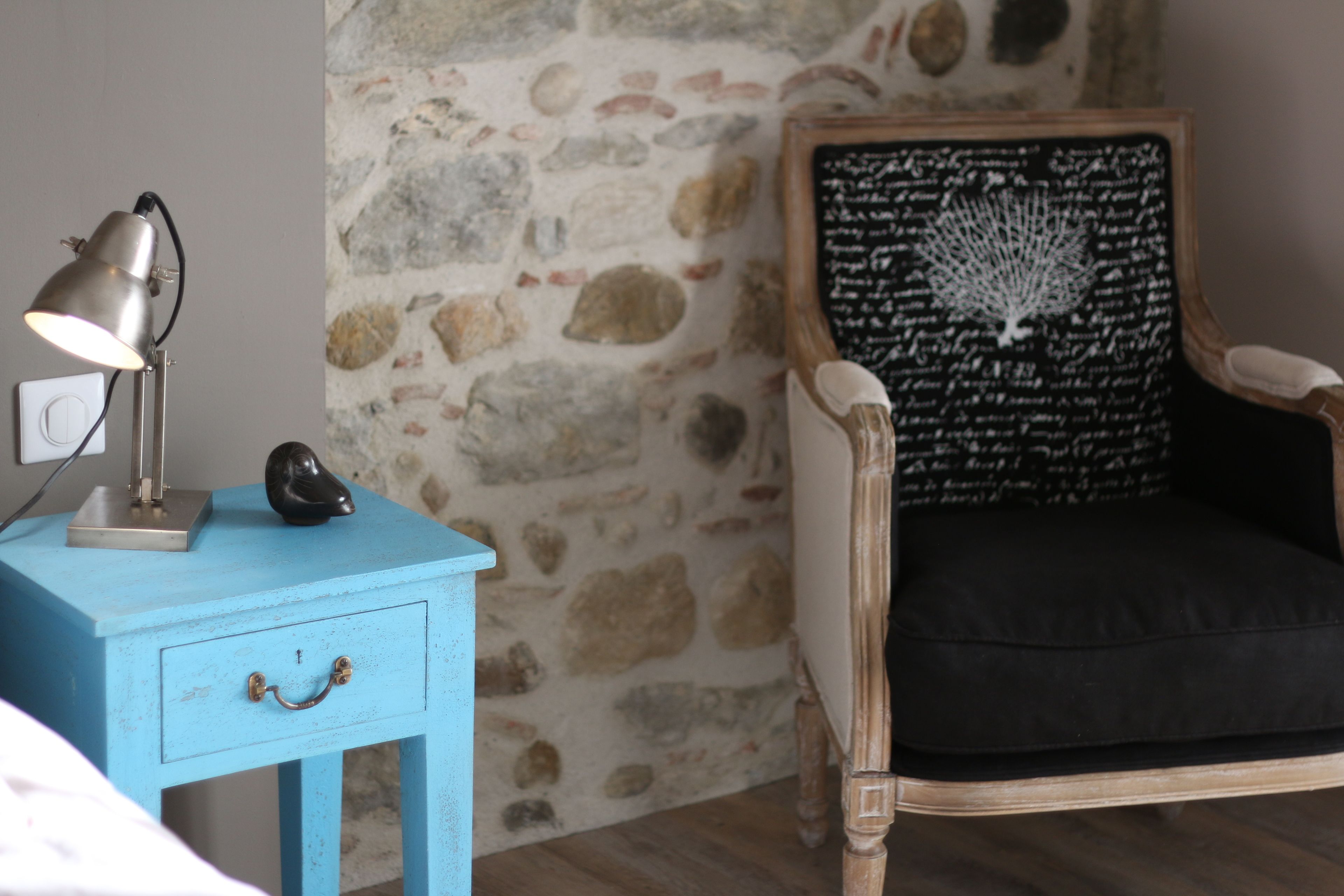 A room with a blue nightstand, a lamp, a decorative object, a chair with a black cushion, and a stone wall.