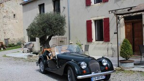 A vintage black convertible car parked in front of a building with red shutters and a stone wall.