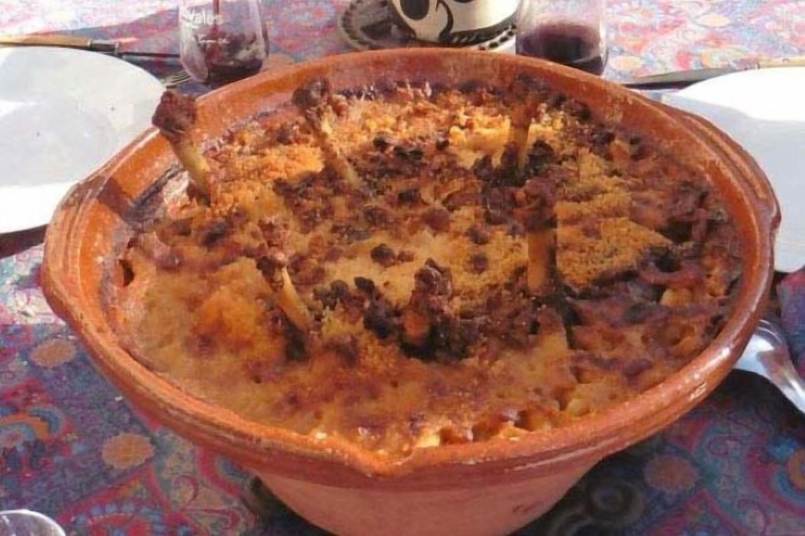 Una olla de barro grande llena de un plato al horno, posiblemente una cazuela, sobre una mesa con mantel estampado.