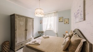 A bedroom with a large wardrobe, a bed with pillows, a bedside table with a lamp, and a window with sheer curtains.