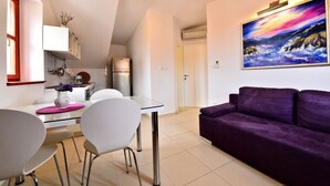 Ein modernes Wohnzimmer mit einem violetten Sofa, einem Glastisch mit Stühlen und einer Küchenzeile. An der Wand hängt ein Bild.