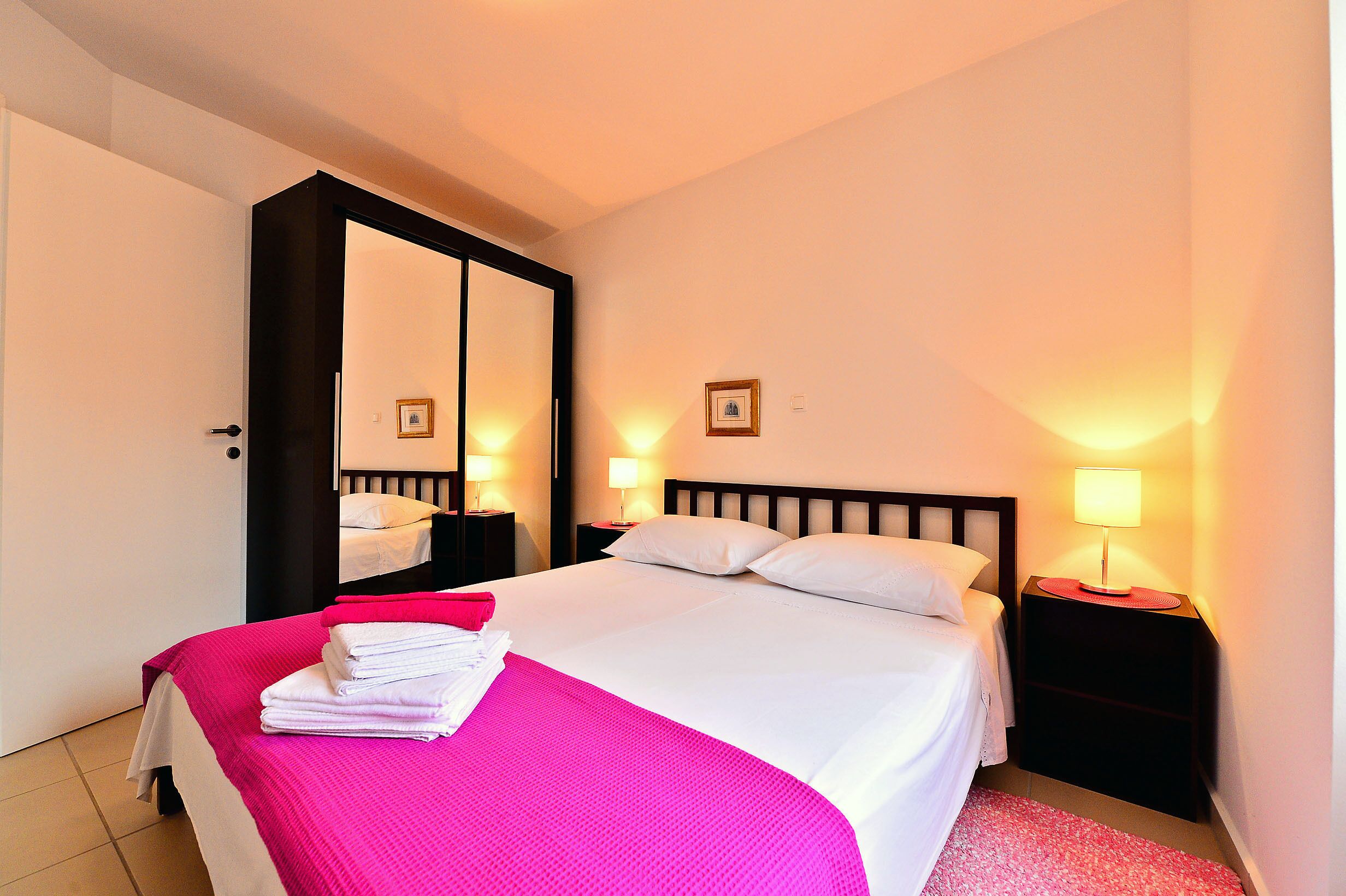 A neatly made bed with a pink blanket, two bedside tables with lamps, a large mirror, and a framed picture on the wall.