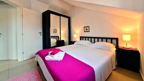 Ein ordentlich gemachtes Bett mit einer rosa Decke, zwei Nachttische mit Lampen, ein großer Spiegel und eine Tür links.