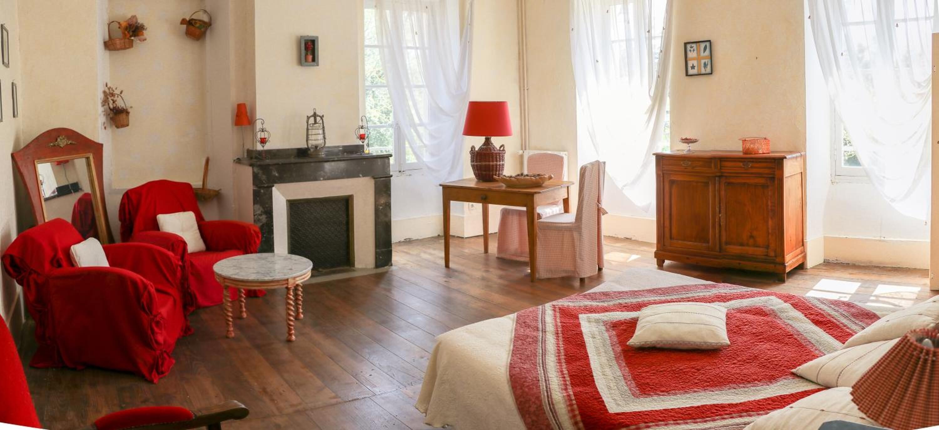 Una habitación acogedora con chimenea, una cama con una manta roja y blanca, un sofá rojo, un armario de madera y una mesita con una lámpara roja.