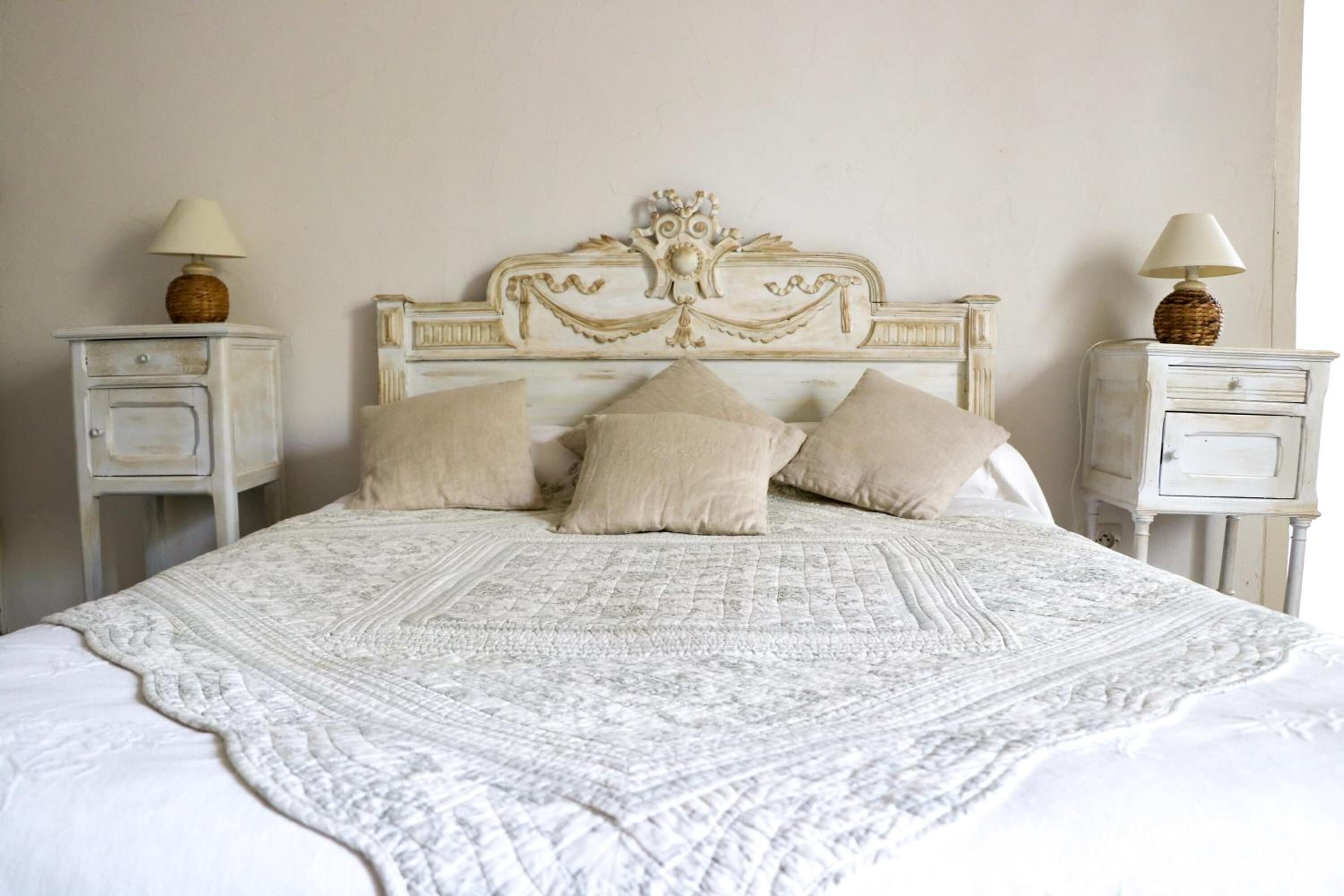 A bed with a white quilt and four beige pillows, flanked by two nightstands with lamps.