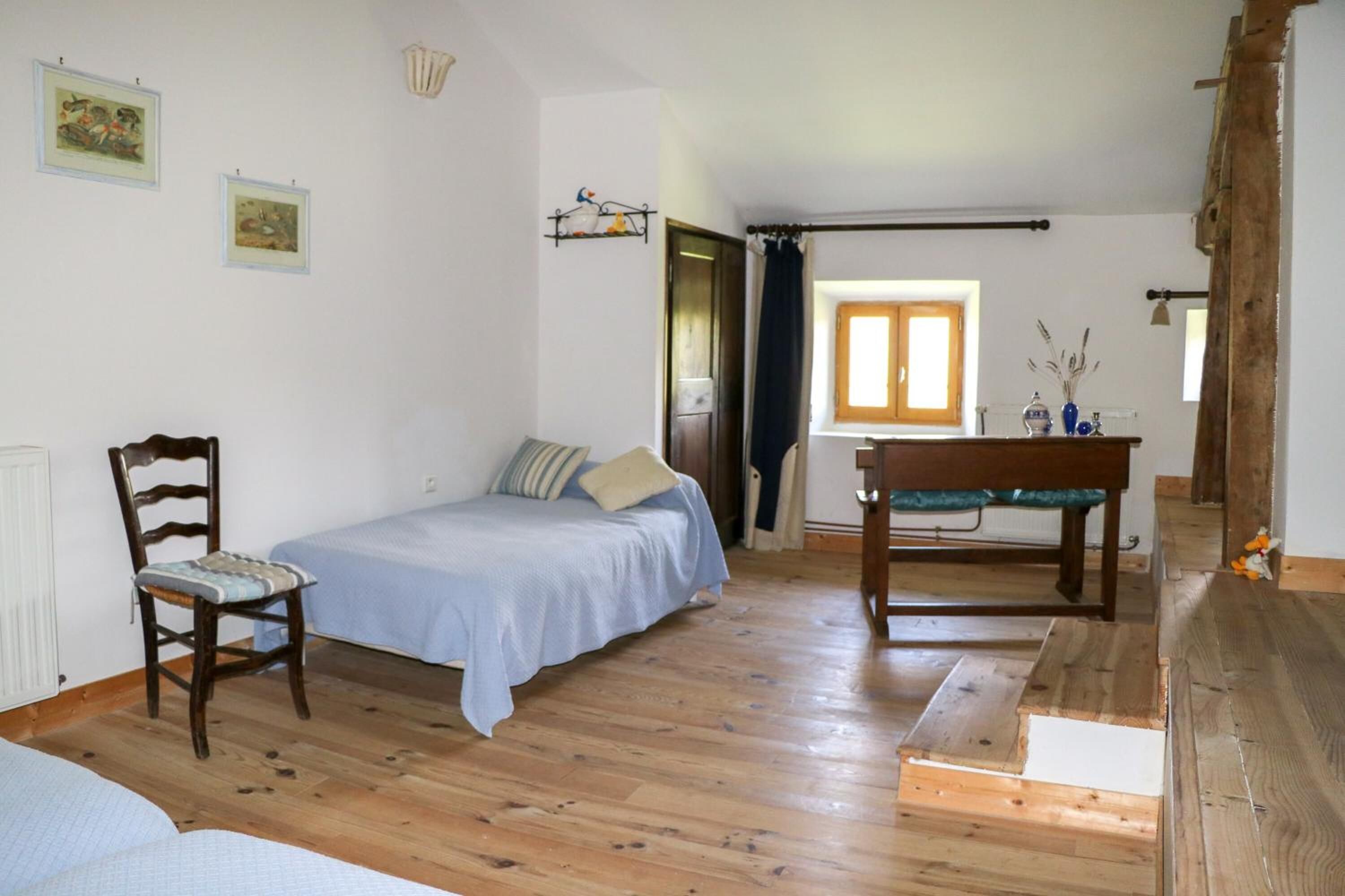 Un dormitori amb llit, una cadira de fusta, una taula petita i una finestra amb cortines.