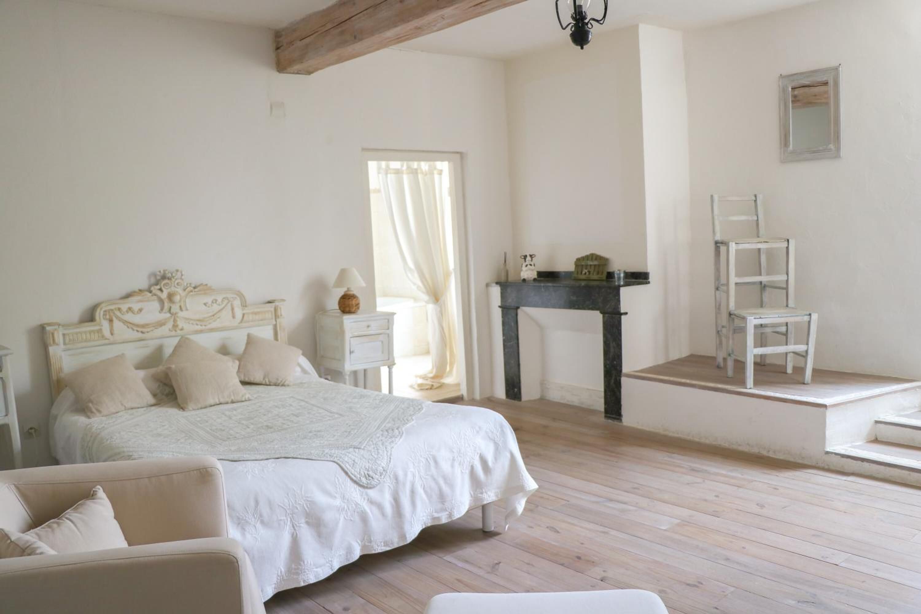 A bedroom with a bed, a chair, a nightstand, a lamp, a fireplace, and a mirror on the wall.