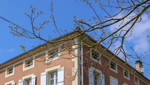 Un edificio de dos plantas con contraventanas blancas, un jardín con macetas de piedra y un cielo azul despejado.
