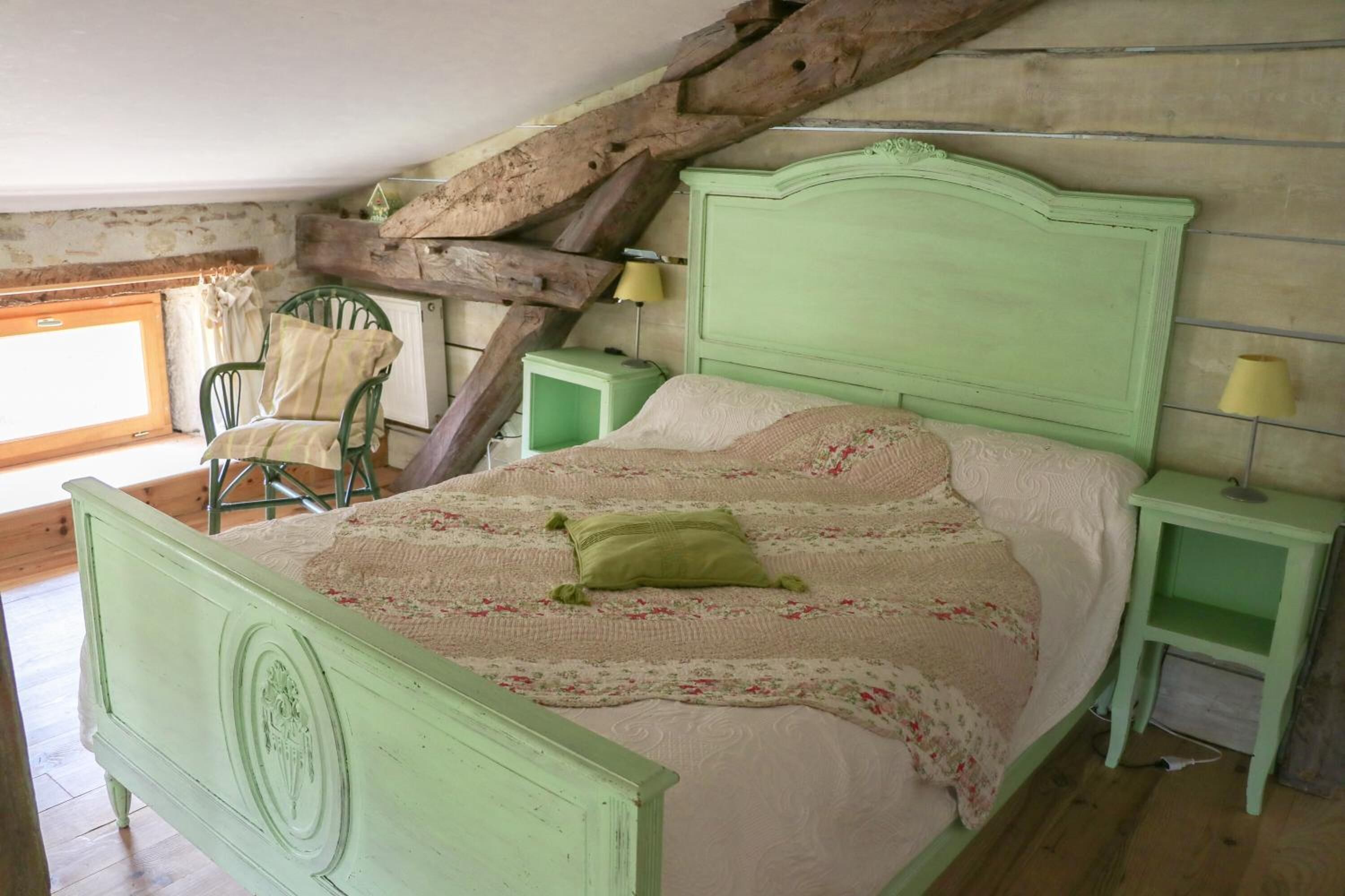 A green-painted bed with a floral bedspread and a single green pillow in a rustic room with exposed wooden beams and a window.