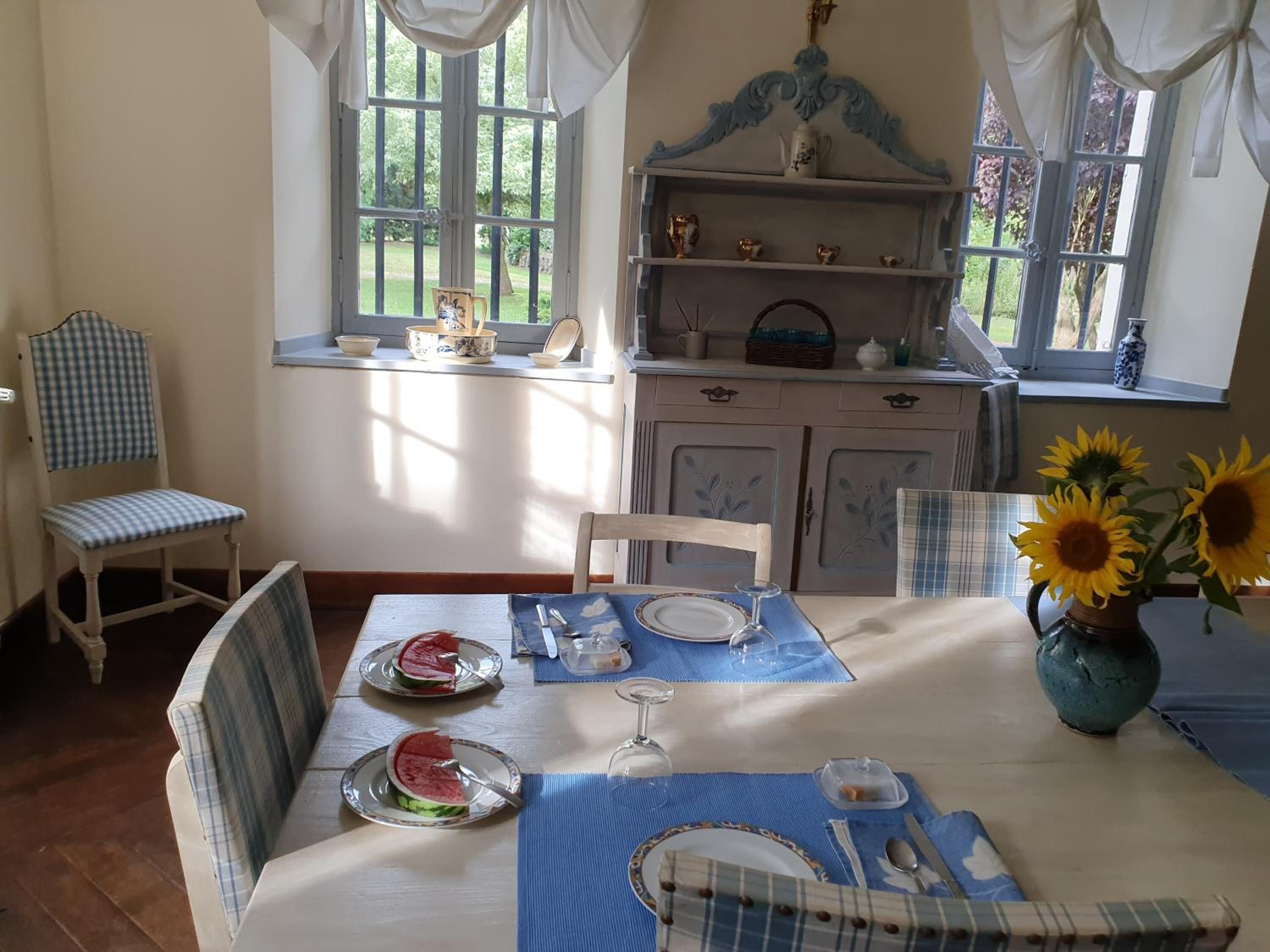A dining area with a table set for two, a window with a view of greenery, and a shelf with decorative items.