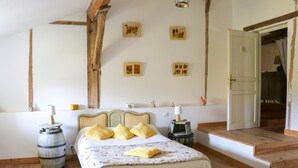 Un dormitorio con cama de madera, una mesita pequeña y una silla. Se ve una puerta que da a otra habitación.