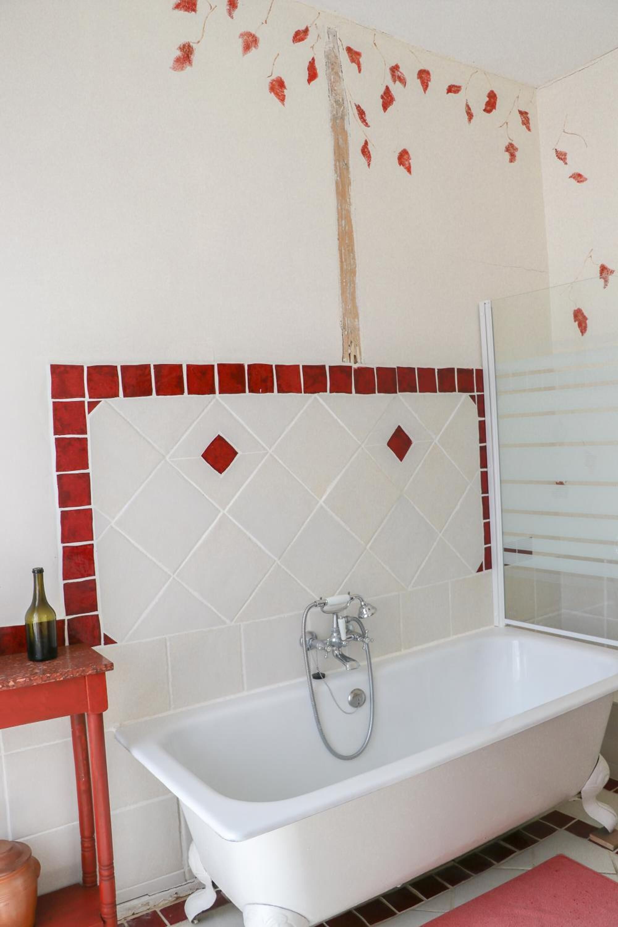 Een badkamer met een wit bad, een wand betegeld in rood en wit, en een klein tafeltje met een fles.
