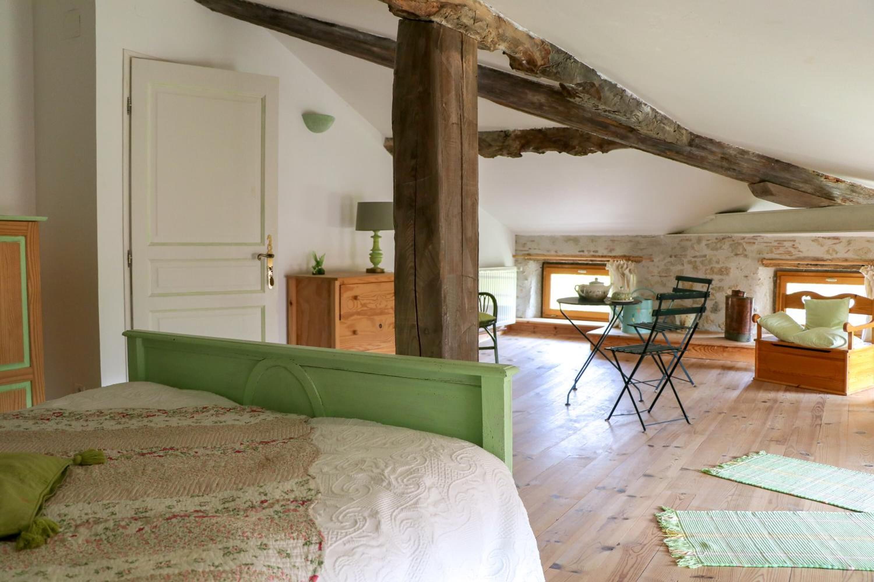 Ein Schlafzimmer mit einem grünen Bett, Holzbalken, einer Essecke mit Tisch und Stühlen und Blick ins Freie.