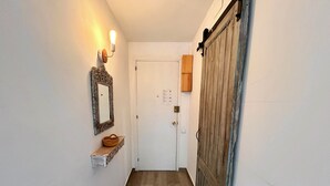 Un pasillo con una puerta blanca, suelo de madera, una lámpara de pared y un espejo.