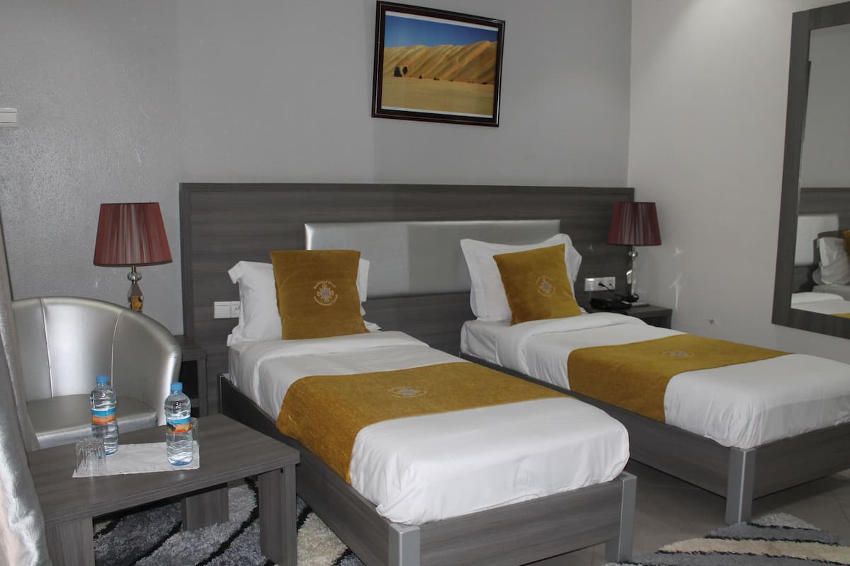 A hotel room with two beds, each with white bedding and gold accents, a desk with a chair, and a nightstand with lamps.
