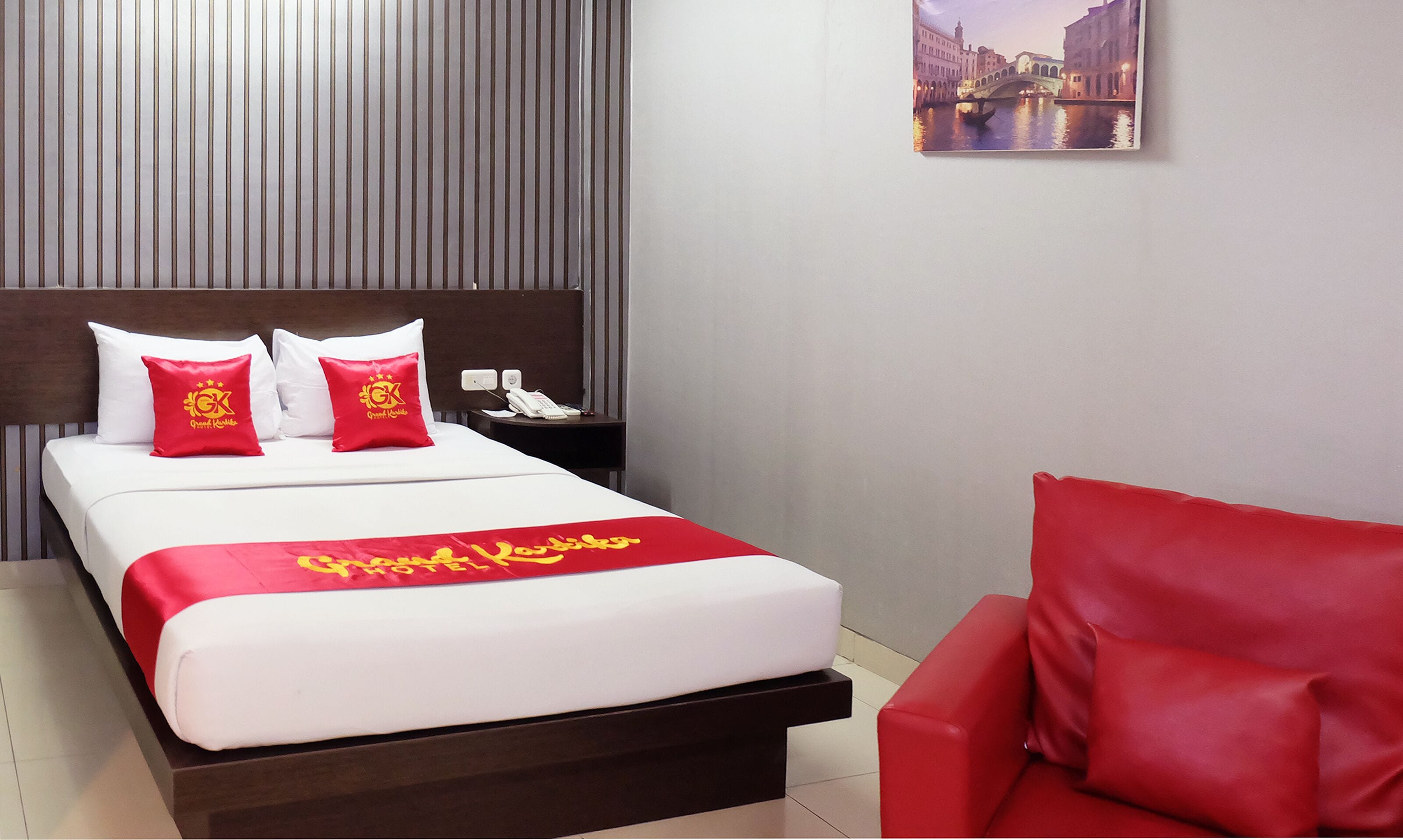 A hotel room with a bed, a nightstand, a red chair, and a wall-mounted picture of a cityscape.
