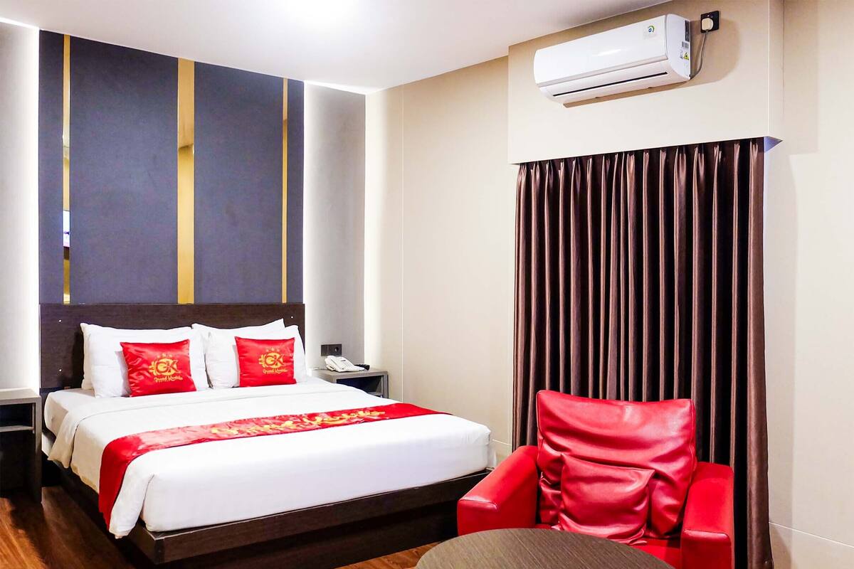 A hotel room with a bed, a red chair, a small table, an air conditioning unit, and a telephone on the nightstand.