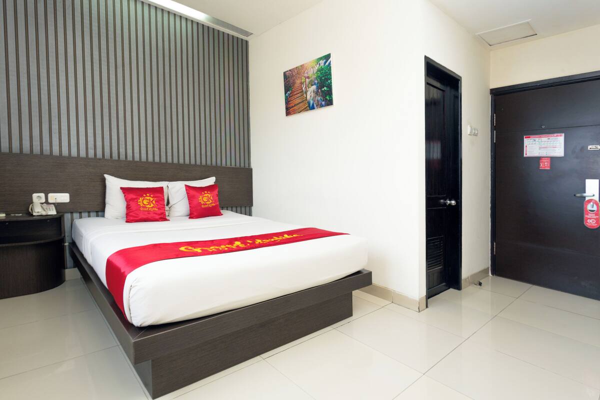 A modern hotel room with a large bed, a bedside table, a wall-mounted artwork, and a door with a fire safety sign.
