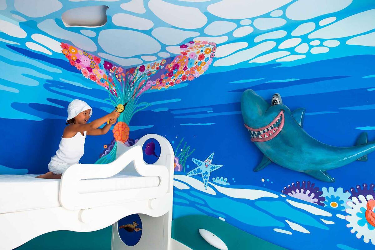 Criança brincando com um brinquedo em uma cama em quarto com um mural de um cenário subaquático.