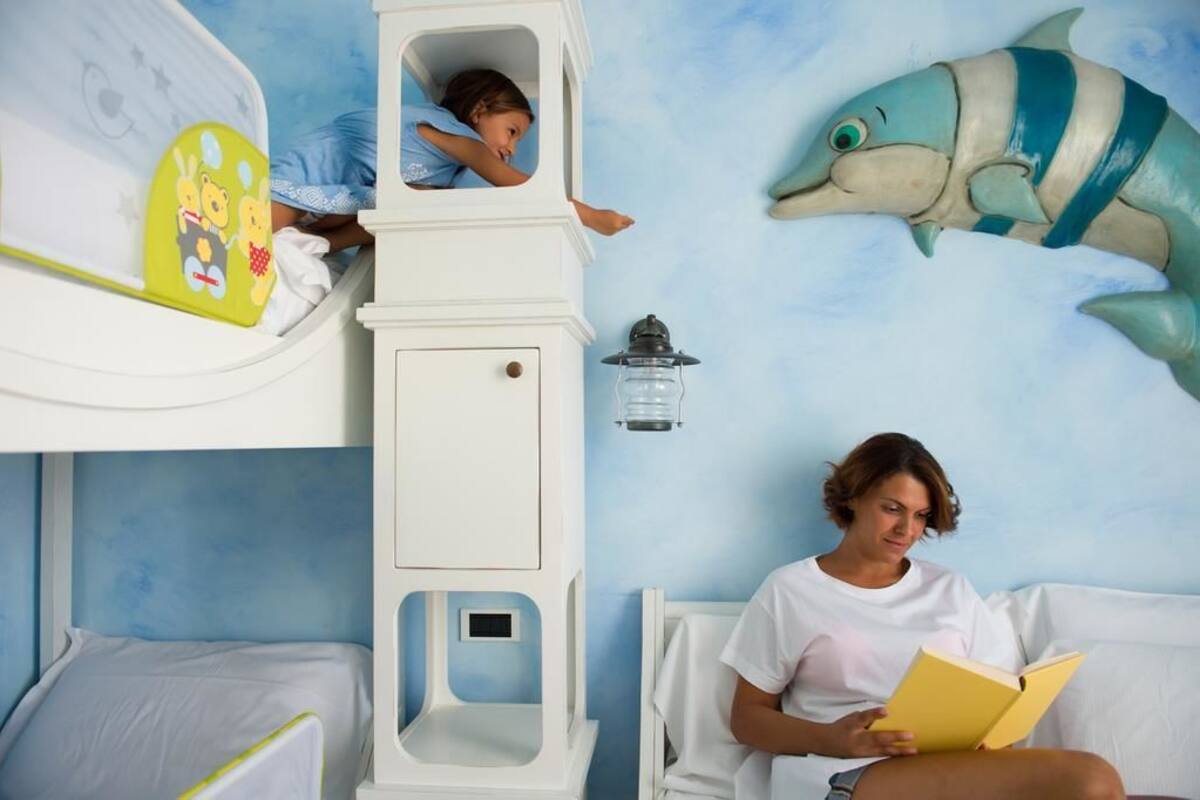 Quarto com beliche, uma criança no beliche, uma mulher lendo e um grande mural de peixes.