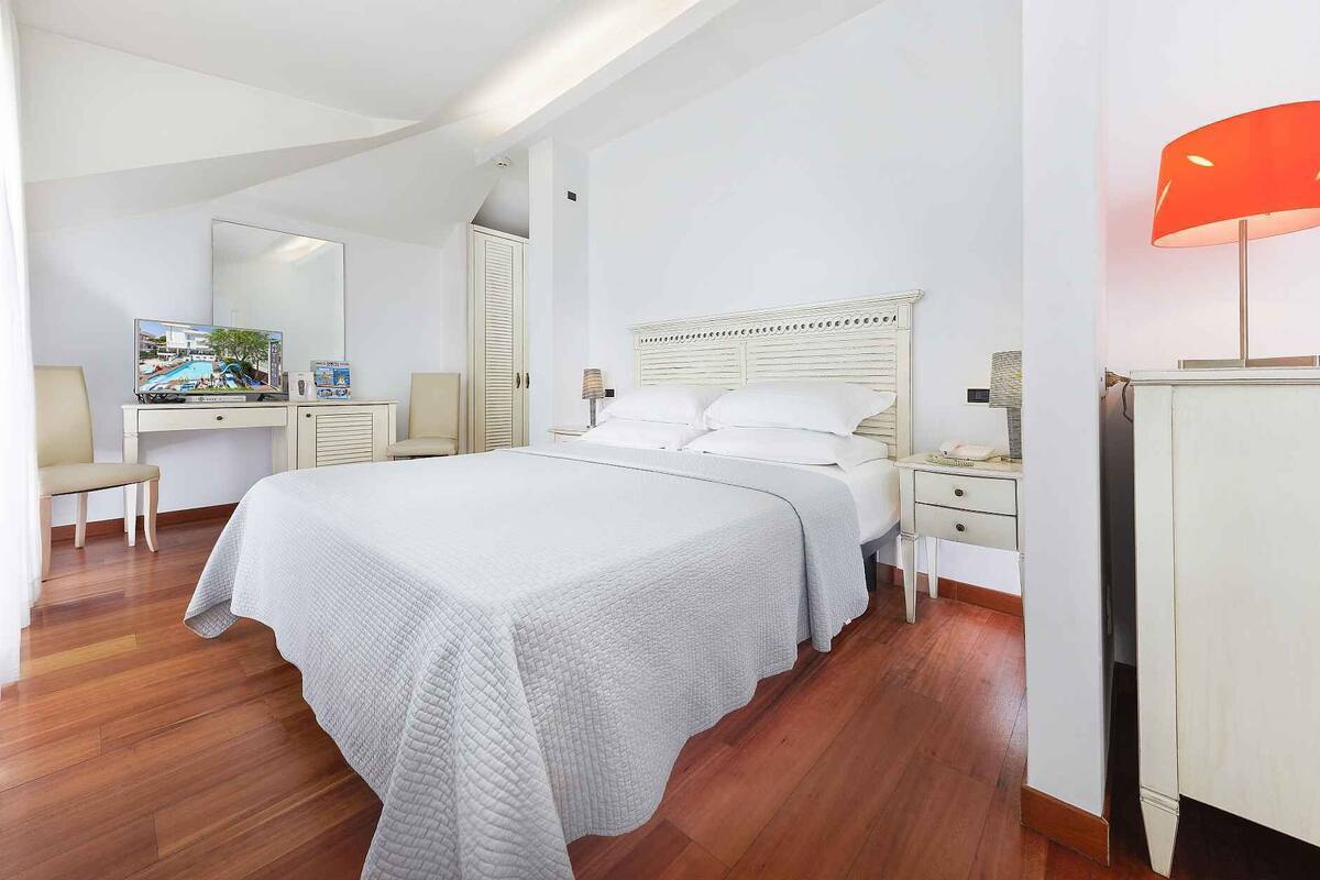 Quarto de hotel com uma cama grande, escrivaninha com televisão, cadeira, guarda-roupa e abajur.