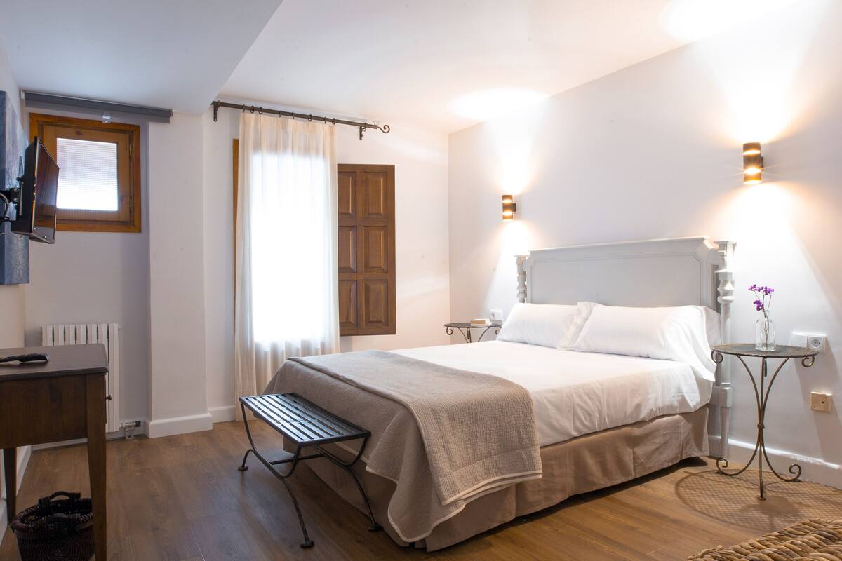 A bedroom with a bed, bedside tables, a bench, a small table with a vase, and a window with curtains.