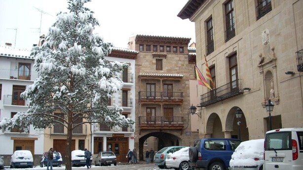 A snow-covered street with parked cars and a building with balconies and an archway. - Hotel Jaime I (Mora de Rubielos)