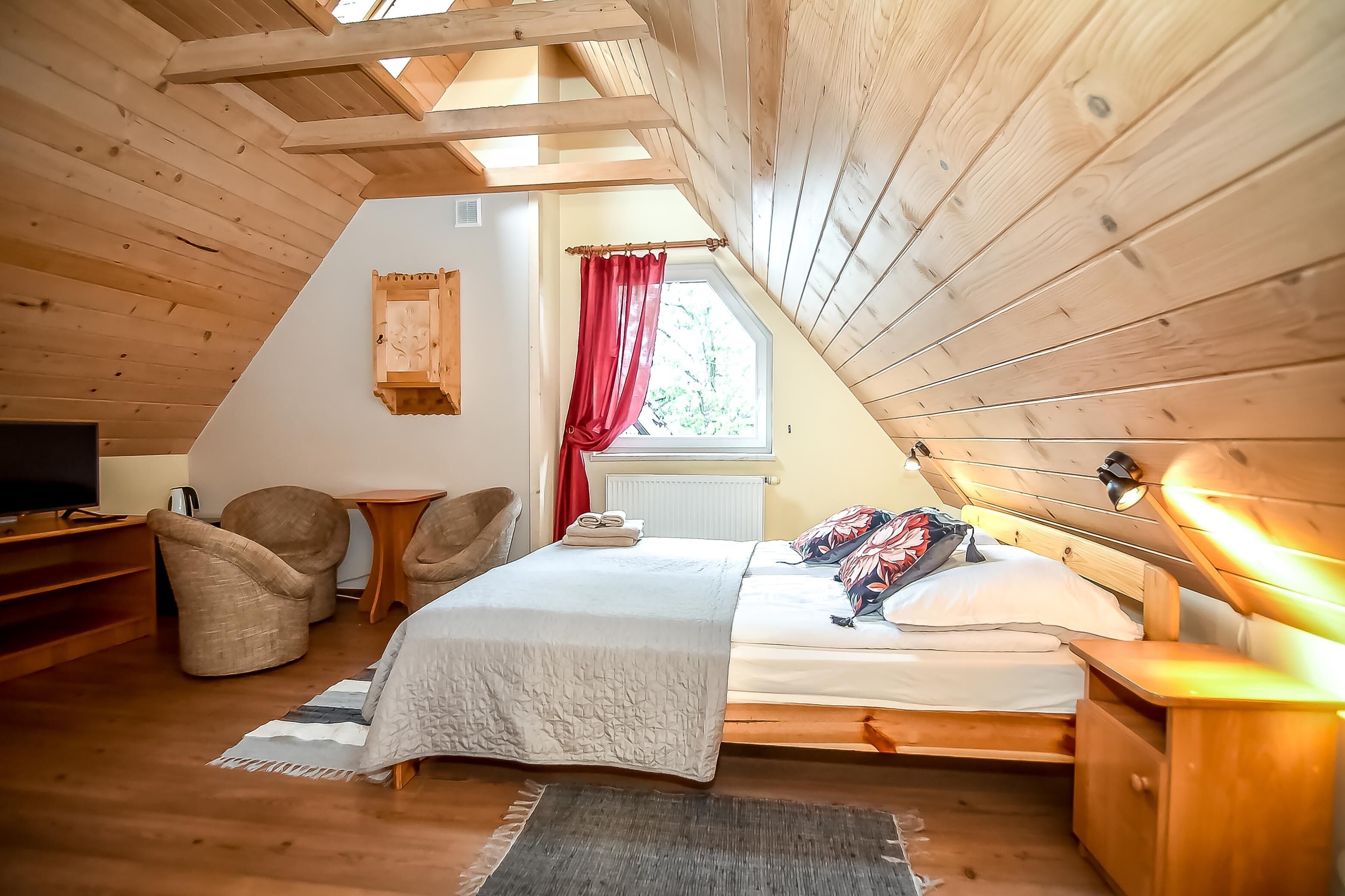 Un dormitorio acogedor en el ático con techo inclinado de madera, una cama con cubrecama blanco, dos sillones, una mesita pequeña y un televisor sobre un aparador de madera.