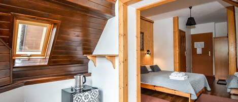 Una habitación acogedora con techo inclinado, una cama con colcha gris, una mesita con un hervidor y una ventana con marco de madera.