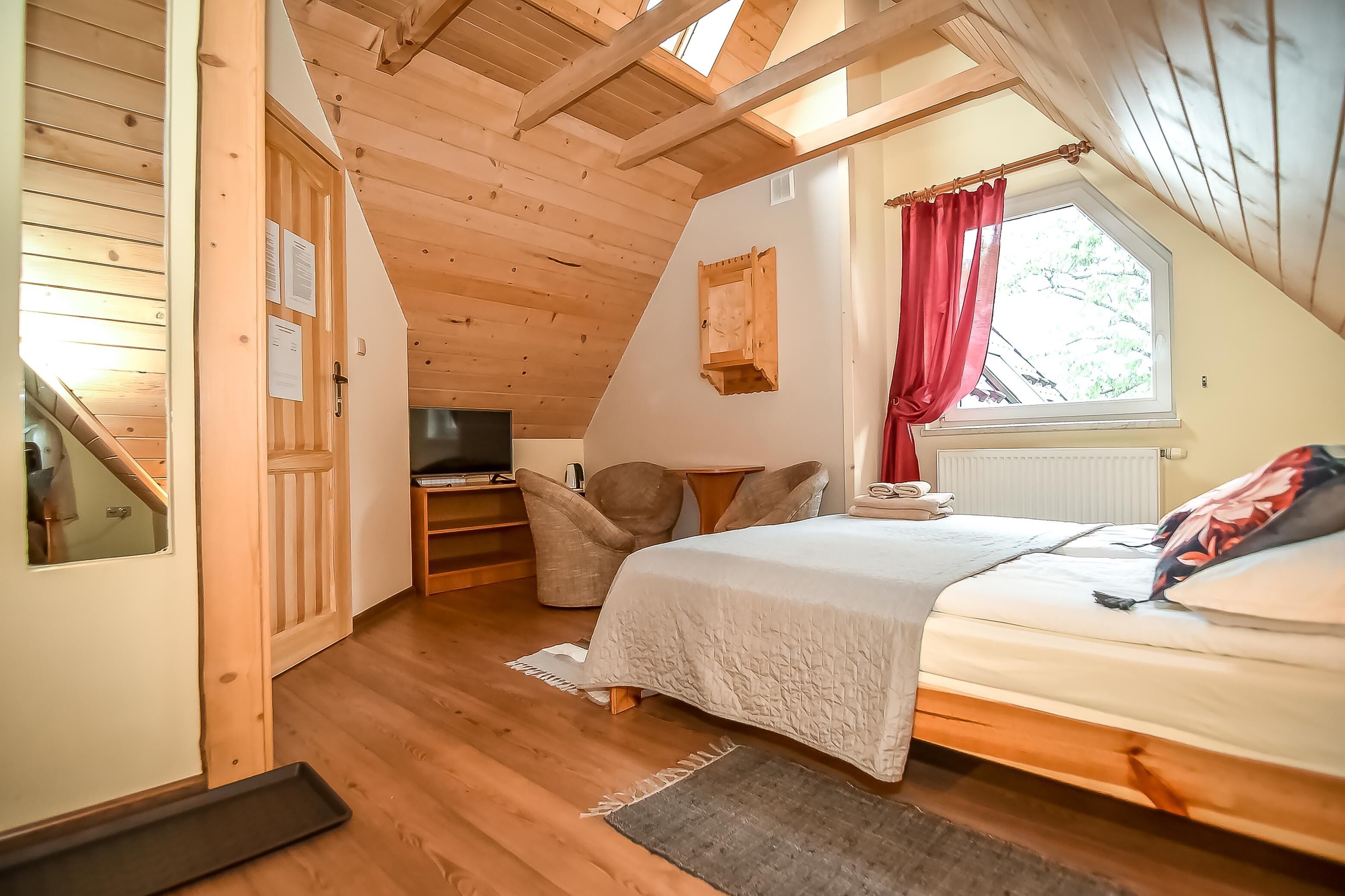 Un dormitorio acogedor en el ático con techo de madera, un ventanal con cortinas rojas, una cama y un televisor en la esquina.