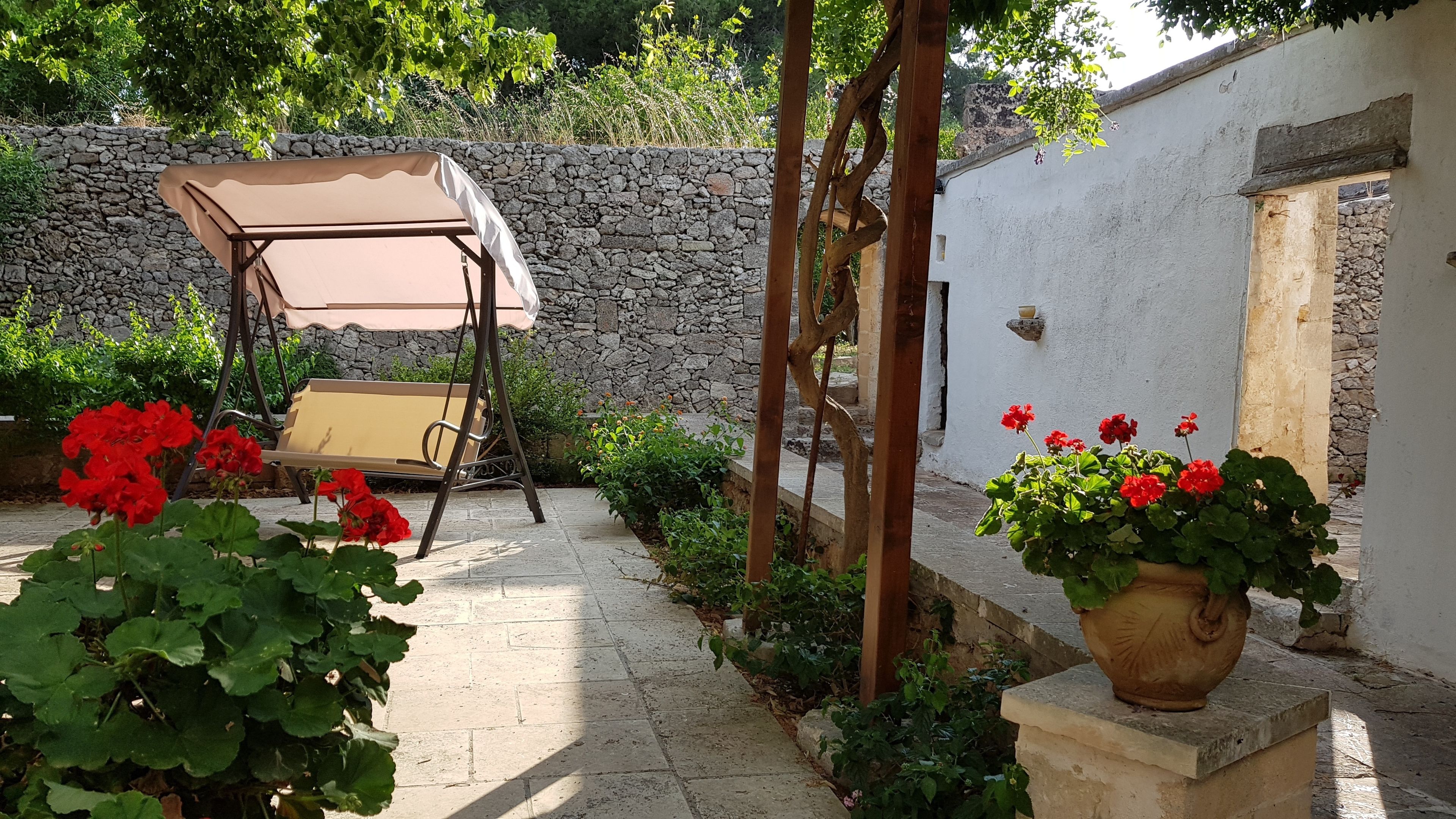 Une terrasse avec un fauteuil suspendu, des géraniums en pot et un mur en pierre.