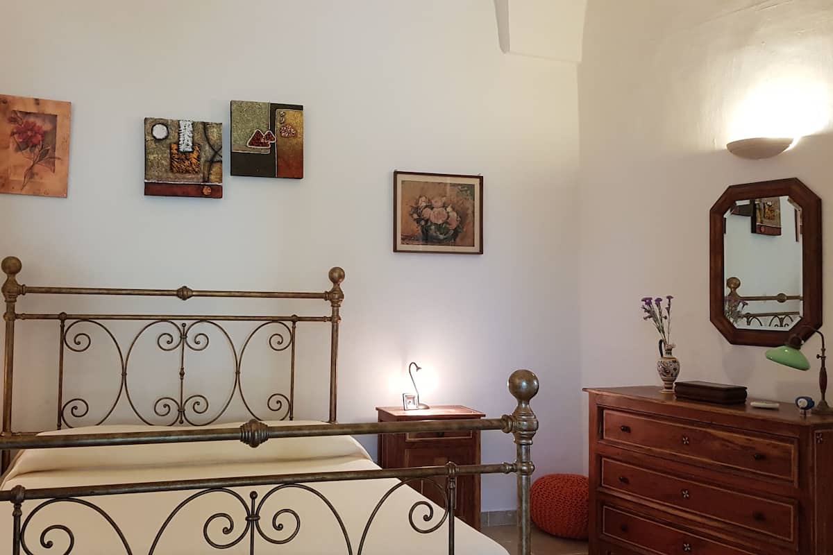 A bedroom with a metal-framed bed, wooden dresser, and a mirror. There are four paintings on the wall.