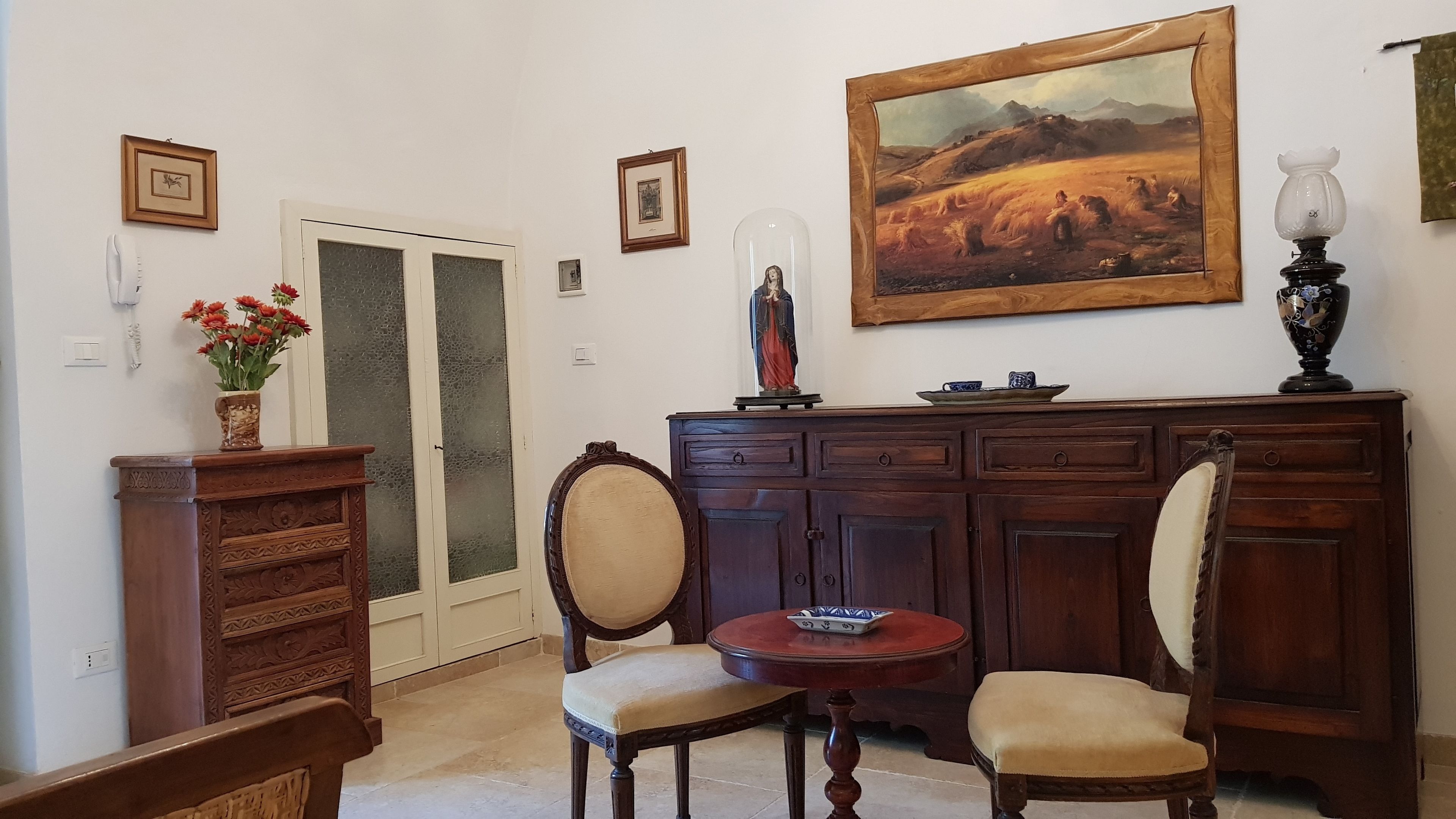 A room with a wooden cabinet, two chairs, a small table, a painting, and a vase with flowers.