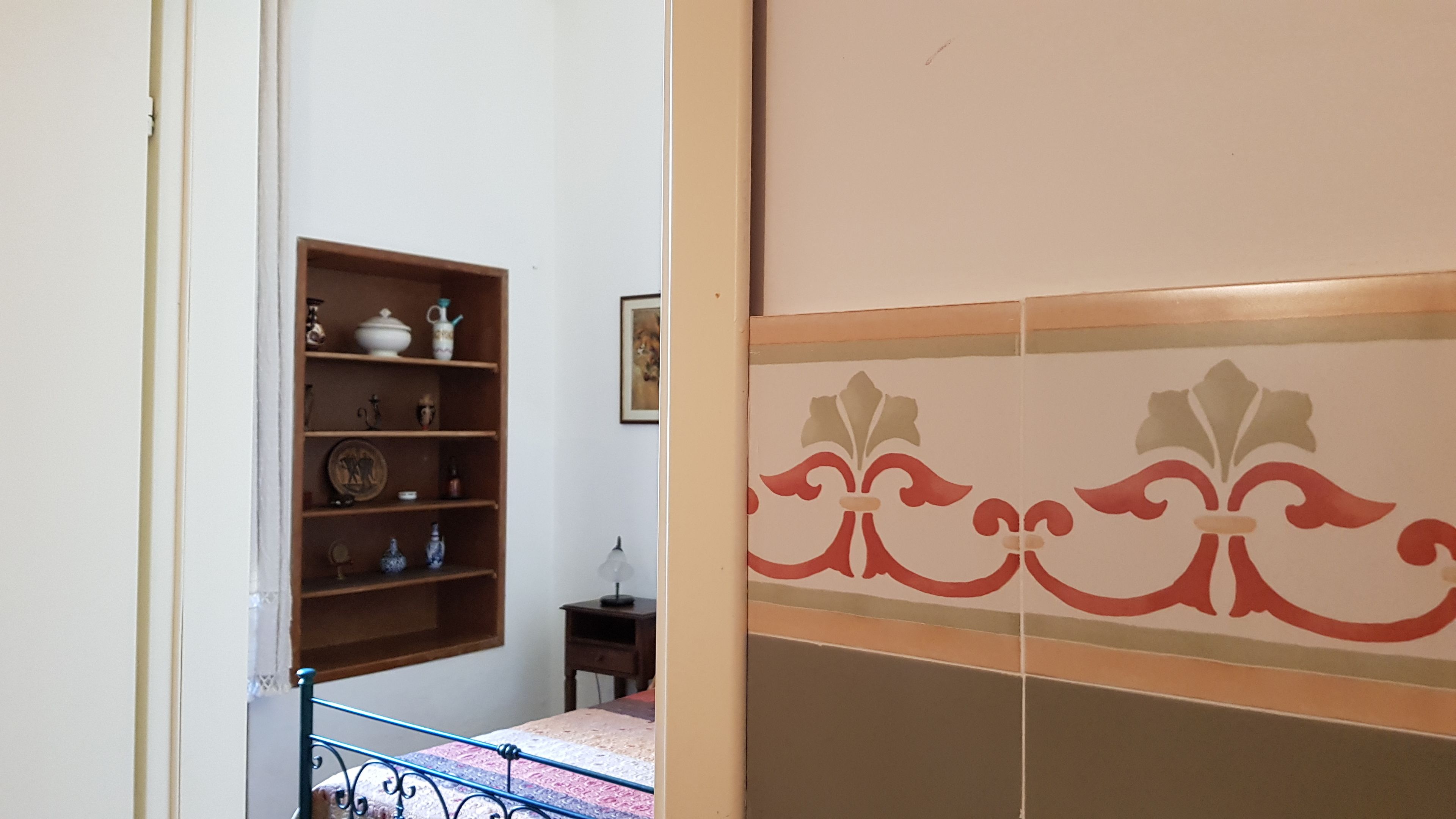 A room with a bed, a wooden shelf with various items, a small table, and a decorative wall panel.
