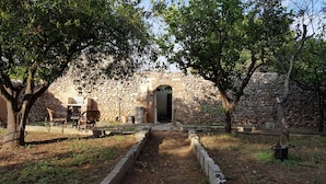 A stone building with a central doorway, surrounded by trees and a dirt path leading to a seating area with chairs and a table. - B&B Le Due Corti (Martignano)