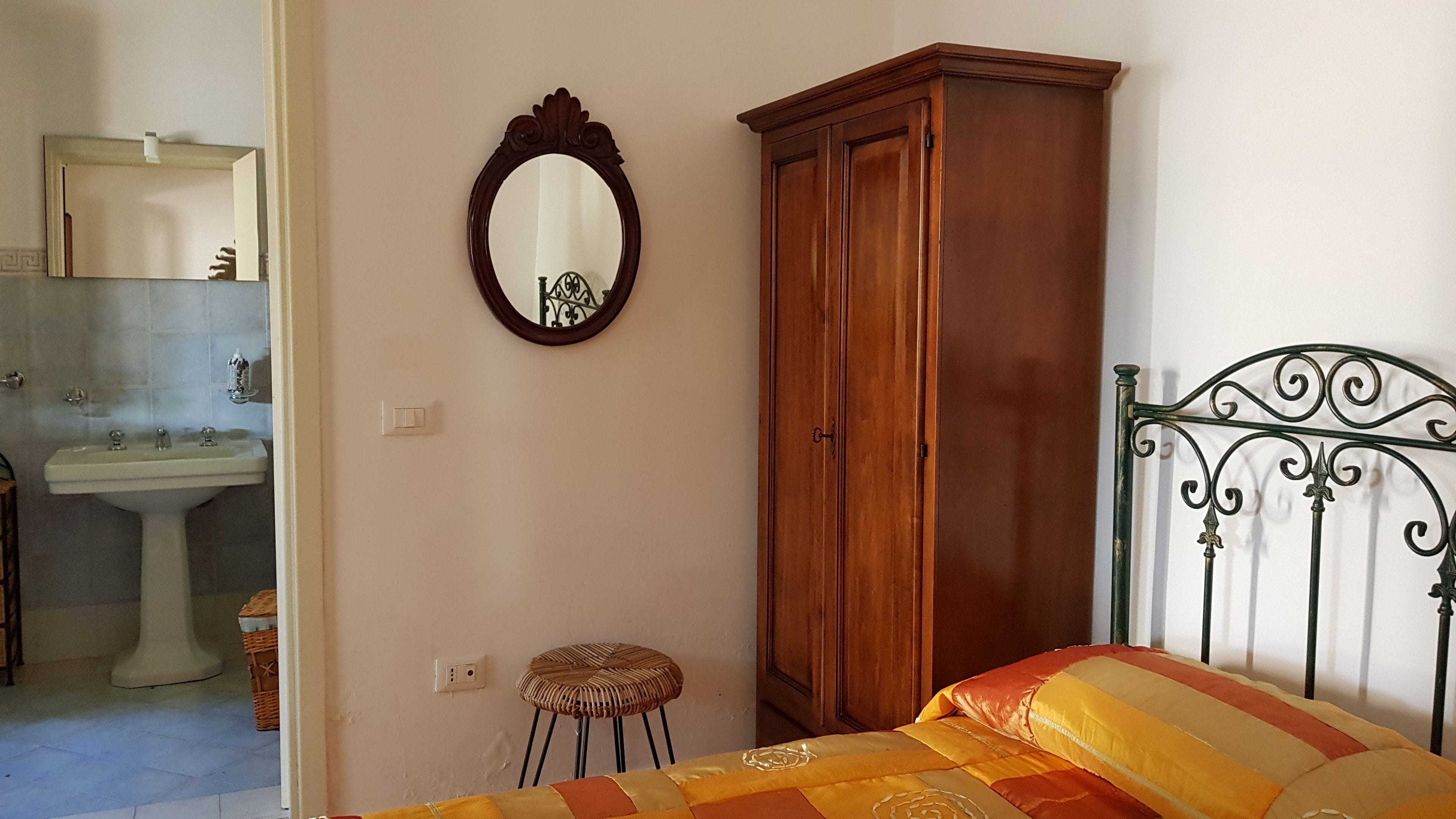 A bedroom with a wooden wardrobe, a round mirror, a bed with striped bedding, a small stool, and a bathroom with a sink and mirror.
