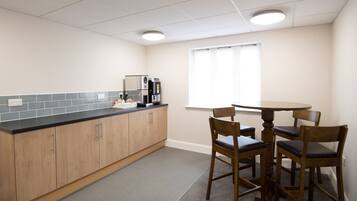 A small, clean kitchen with wooden cabinets, a black countertop, and a coffee machine. There is a round table with four chairs and a window with blinds.