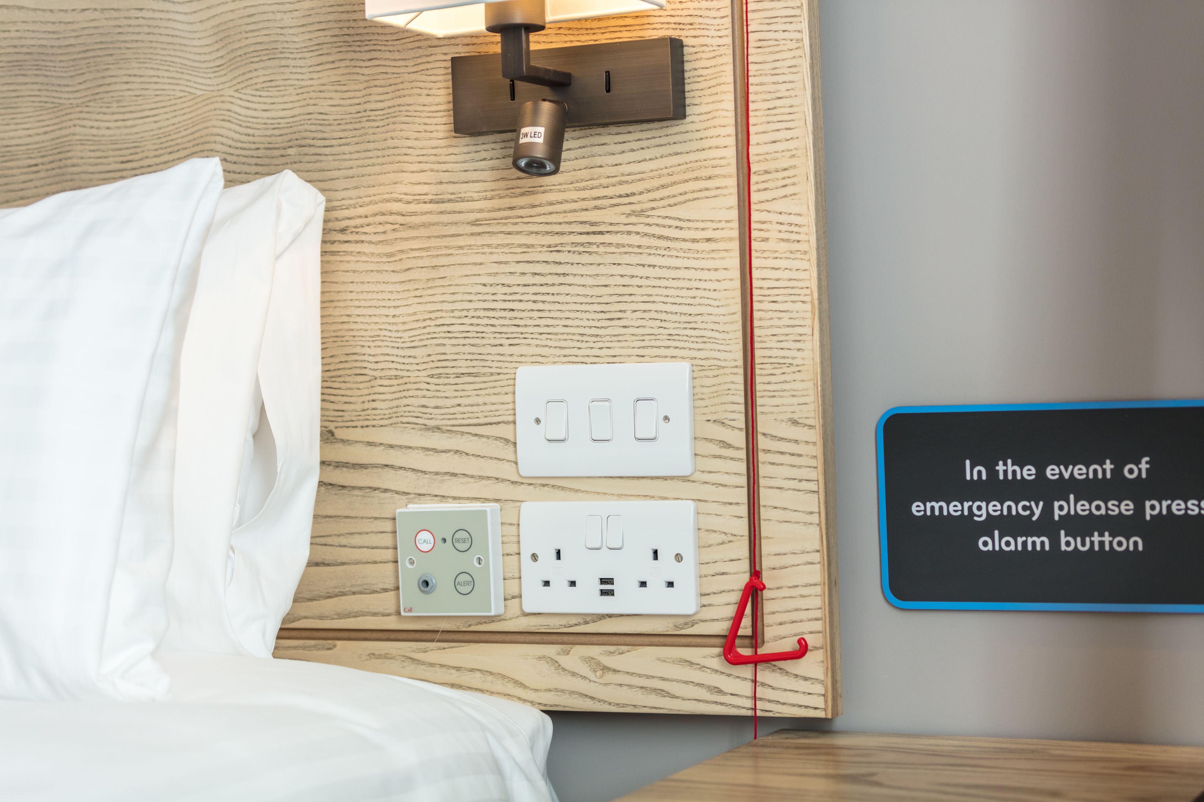 Habitación de hotel con cama, una lámpara de pared, una cámara de seguridad y un botón de alarma de emergencia.