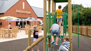 A playground with a wooden climbing structure, a slide, and a brick building labeled "PAISLEY PEAR" in the background. - Paisley Pear, Brackley by Marston's Inns (Brackley)