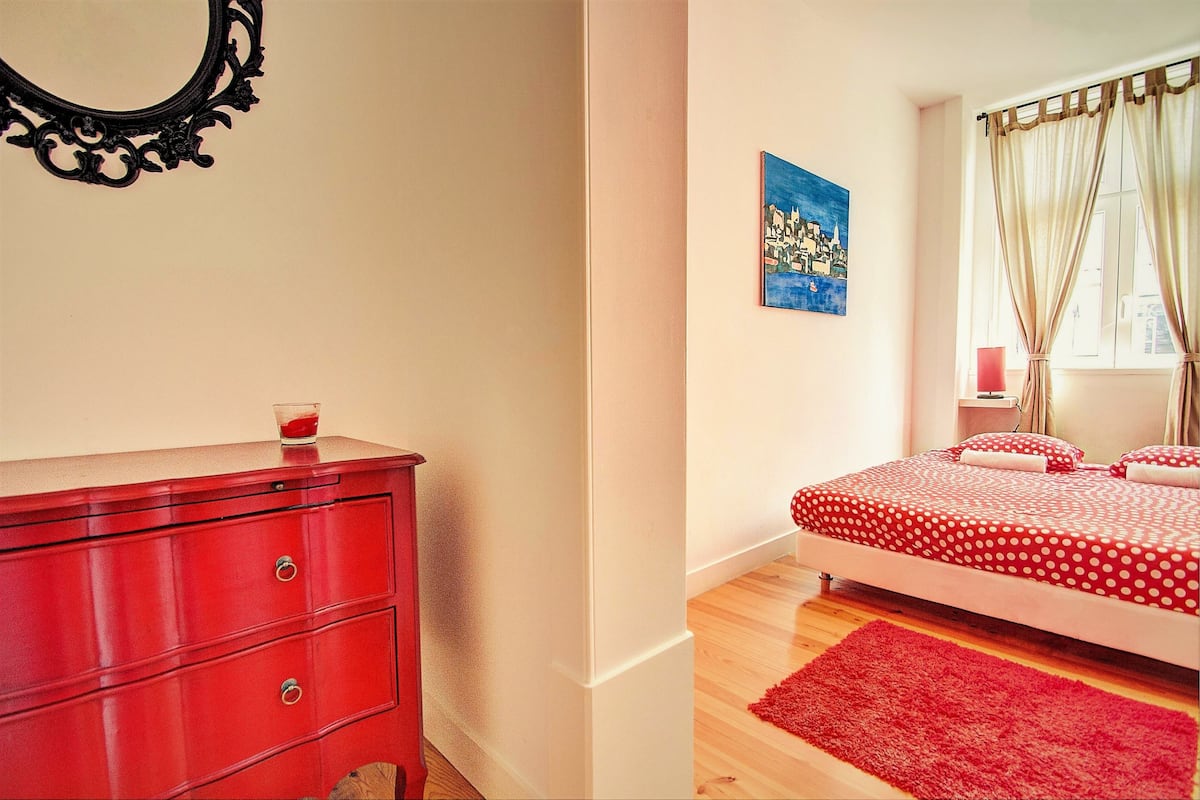 Une chambre avec une commode rouge, un lit recouvert d’une couette à pois rouges et blancs, un tapis rouge, un tableau accroché au mur et une fenêtre avec des rideaux.