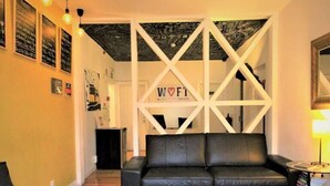 A modern living room with a black leather sofa, a grey armchair, and a colorful striped rug. There is a white geometric wall structure with the letters "W O F T" and a small table with a lamp.