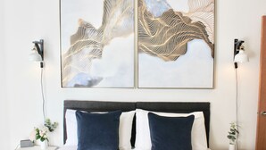 A neatly made bed with two pillows, a bedside table with a lamp, and two wall-mounted paintings. - Tincroft Cottage Luxury Coastal Living (Truro)