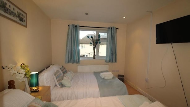 A bedroom with two beds, a window with curtains, a flat-screen TV, and a bedside table with a lamp and flowers. - Atchison Gardens (Derby)