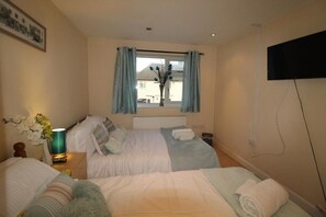 A bedroom with two beds, a window with curtains, a flat-screen TV, and a bedside table with a lamp and flowers. - Atchison Gardens (Derby)