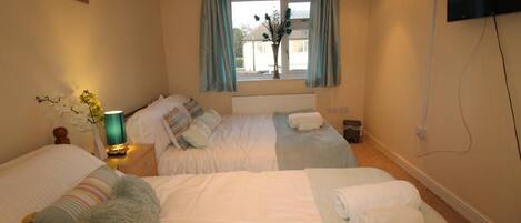 A hotel room with two beds, a window with curtains, a bedside table with a lamp, and a television on the wall.