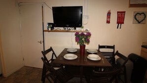 A dining area with a table set for four, a television mounted above, and a small shelf with a plant and a fire extinguisher. - Atchison Gardens (Derby)