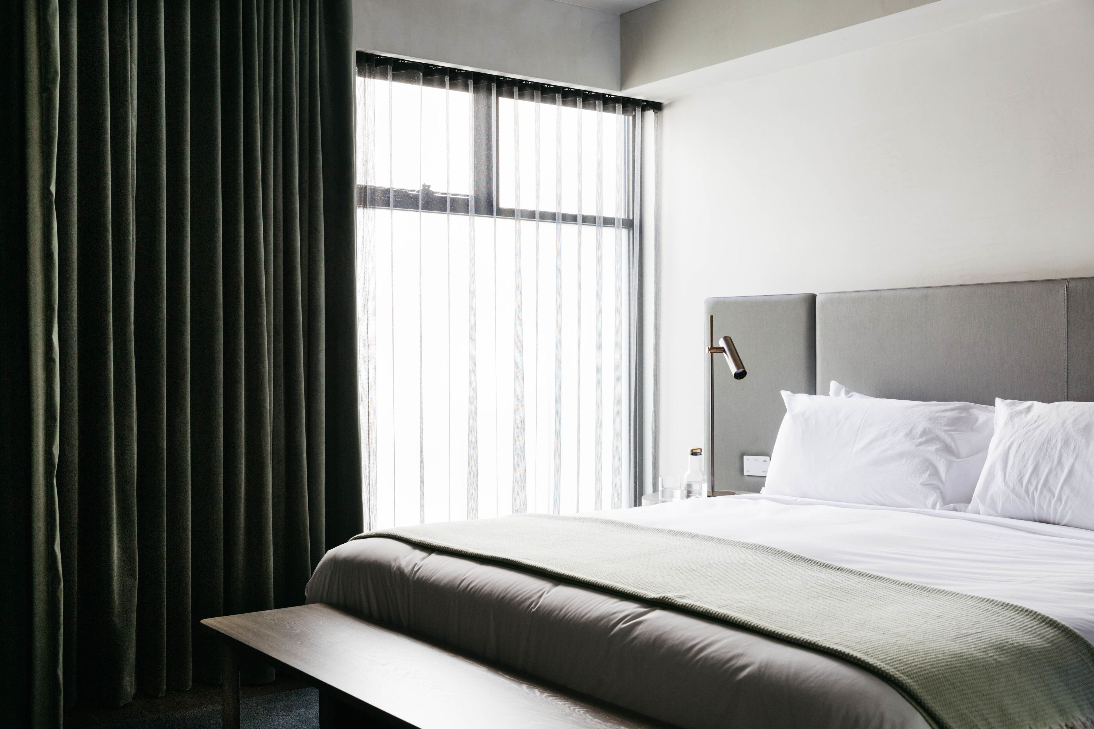 A modern hotel room with a large bed, a bench, and a window with sheer curtains.